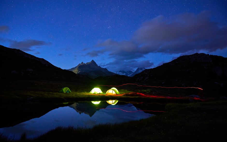 Belle scène de camping nocturne avec des tentes sous un ciel étoilé et des reflets de montagnes dans un lac.