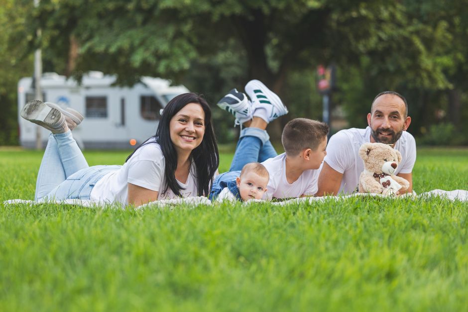 Famille heureuse allongée sur l'herbe avec un camping-car en arrière-plan, profitant d'un pique-nique en plein air sous le soleil.